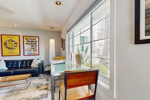 A modern living room with a glass-top dining table, a leather sofa, and a large window with a view of greenery. - Updated modern condo w/ shared pool & mountain views - near downtown (Palm Springs)