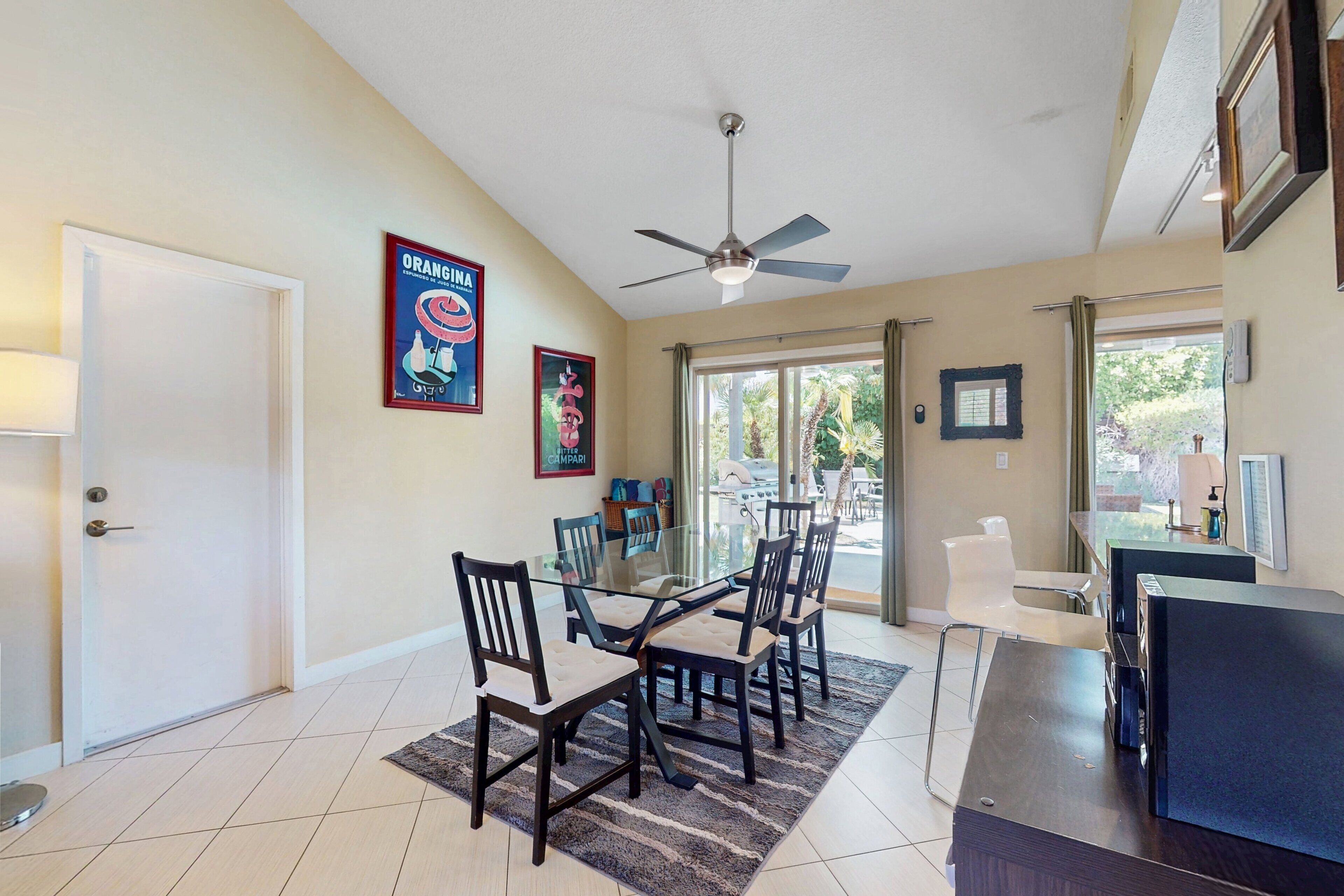 A dining area with a glass table, four chairs, a ceiling fan, and a view of the outdoors.