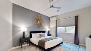 Ein Schlafzimmer mit einem Bett, Nachttischen, einem Deckenventilator, einem großen Fenster mit Vorhängen und einem gemusterten Teppich.
