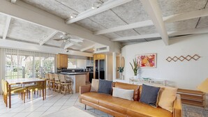 A spacious living area with a sofa, dining table, and chairs. A kitchen is visible in the background with a window overlooking greenery. - Dog-friendly mountain view condo with pool, grill, open layout, AC, & internet (Palm Desert)