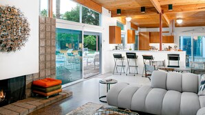 A modern living room with a fireplace, a grey sofa, and a dining area with bar stools. - Elegant, mid-century Alexander butterfly home w/ pool & gas grill (Palm Springs)