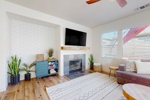 A living room with a fireplace, a flat-screen TV mounted above it, a blue cabinet with decorative items, and a large patterned rug. - Serene home with pool spa, gas fireplace, AC, pool and golf access - dogs ok (Indio)