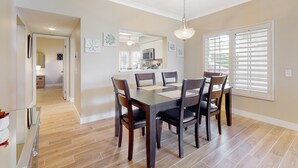 Un coin repas avec une table et des chaises en bois, un luminaire suspendu et, en arrière-plan, une vue sur la cuisine.
