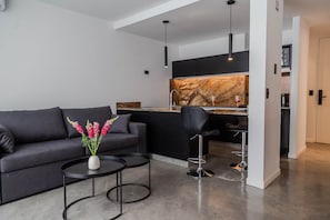 A modern living room with a sofa, a coffee table, a vase of flowers, a kitchen area with a marble backsplash, and a bar stool. - Estudio Moderno & Amplio en Palermo (Buenos Aires)