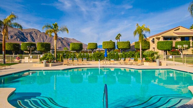 A swimming pool with lounge chairs and palm trees, surrounded by a fence and a mountain in the background. - Dog-friendly condo w/ two shared outdoor pools, spas & tennis court (Palm Springs)