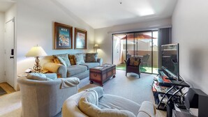A spacious living room with a grey sofa, a coffee table, and a TV. There are two framed pictures on the wall and a lamp on the floor. - Dog-friendly condo w/ two shared outdoor pools, spas & tennis court (Palm Springs)