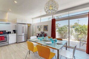 A modern kitchen with stainless steel appliances, a dining area with a glass table and yellow chairs, and a large window with a view of greenery. - Iconic, Wexler-designed, mid-century home w/ 50-foot pool & attached spa (Palm Springs)