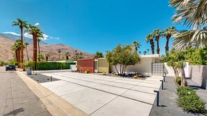 Un área residencial moderna con un camino de entrada que conduce a un acceso con portón, rodeada de palmeras y una montaña al fondo.