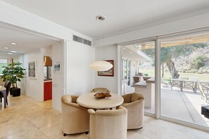 A modern dining area with a round table and beige chairs, a pendant light, and a view of an outdoor patio with seating. - Exquisite retreat with pool, spa, fireplace, golf & mountain view, dog-friendly! (Indian Wells)