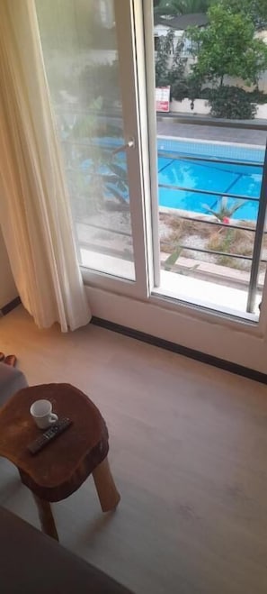 Une pièce dotée d’une grande fenêtre donnant sur une piscine, d’un tabouret en bois avec une télécommande, et d’un rideau blanc.