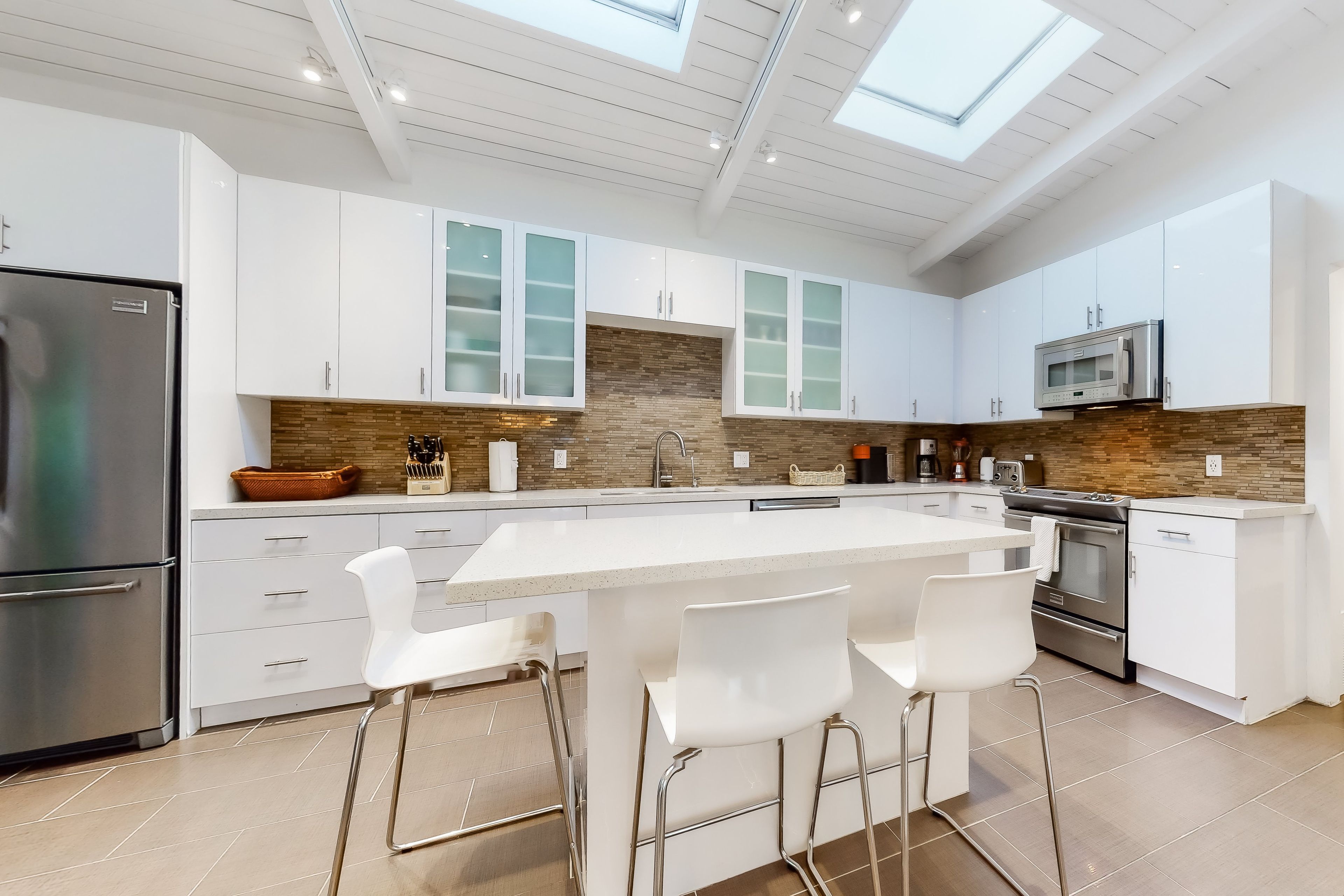 Una cocina moderna con armarios blancos, una isla central con taburetes altos, un refrigerador de acero inoxidable y un microondas.