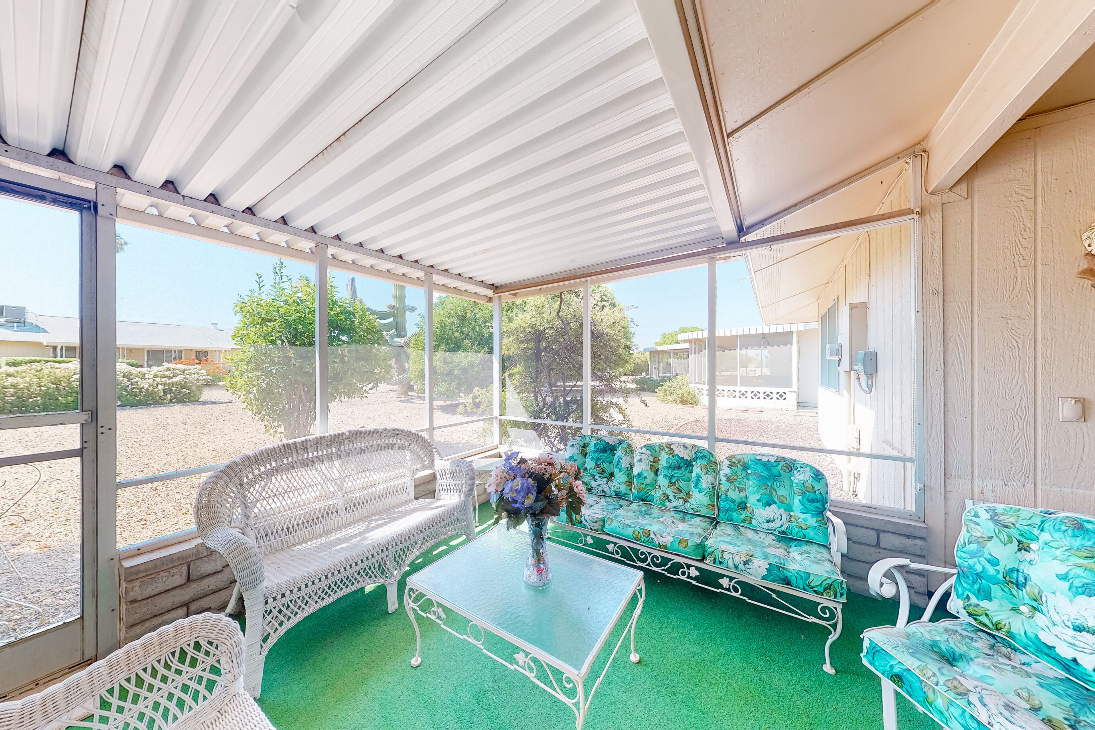 A covered patio with wicker furniture and a glass table, overlooking a garden and buildings.