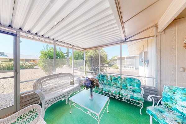 A covered patio with wicker furniture and a glass table, overlooking a garden and buildings.