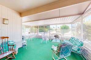 A covered outdoor seating area with green flooring, white wicker furniture, and a view of the outdoors. - Kodiak Cottage | House in Sun City West (Sun City West)