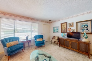 A living room with two armchairs, a glass coffee table, a television, and a cabinet with decorative items. - Kodiak Cottage | House in Sun City West (Sun City West)