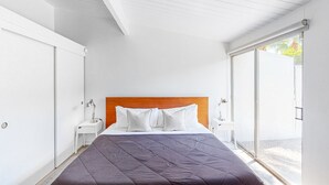 A modern bedroom with a large bed, white walls, and a wooden headboard. - Sophisticated & modern home w/private gas grill, heated outdoor pool, large lawn (Palm Springs)