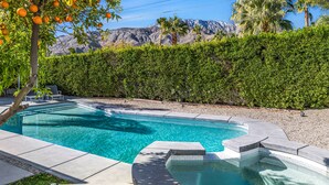 A swimming pool with a curved design, surrounded by a hedge and a mountain backdrop. - Sophisticated & modern home w/private gas grill, heated outdoor pool, large lawn (Palm Springs)