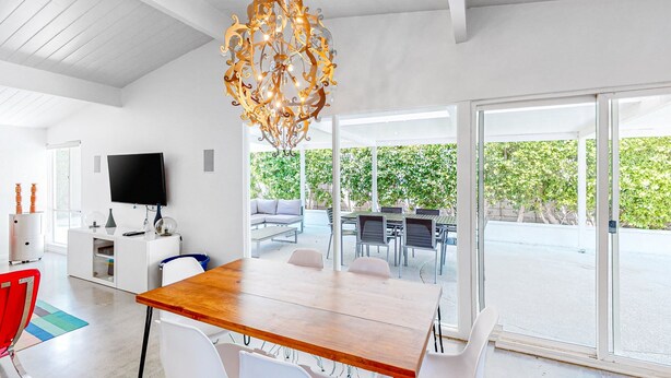A modern dining area with a wooden table and white chairs, a chandelier, and a TV on the wall. - Sophisticated & modern home w/private gas grill, heated outdoor pool, large lawn (Palm Springs)