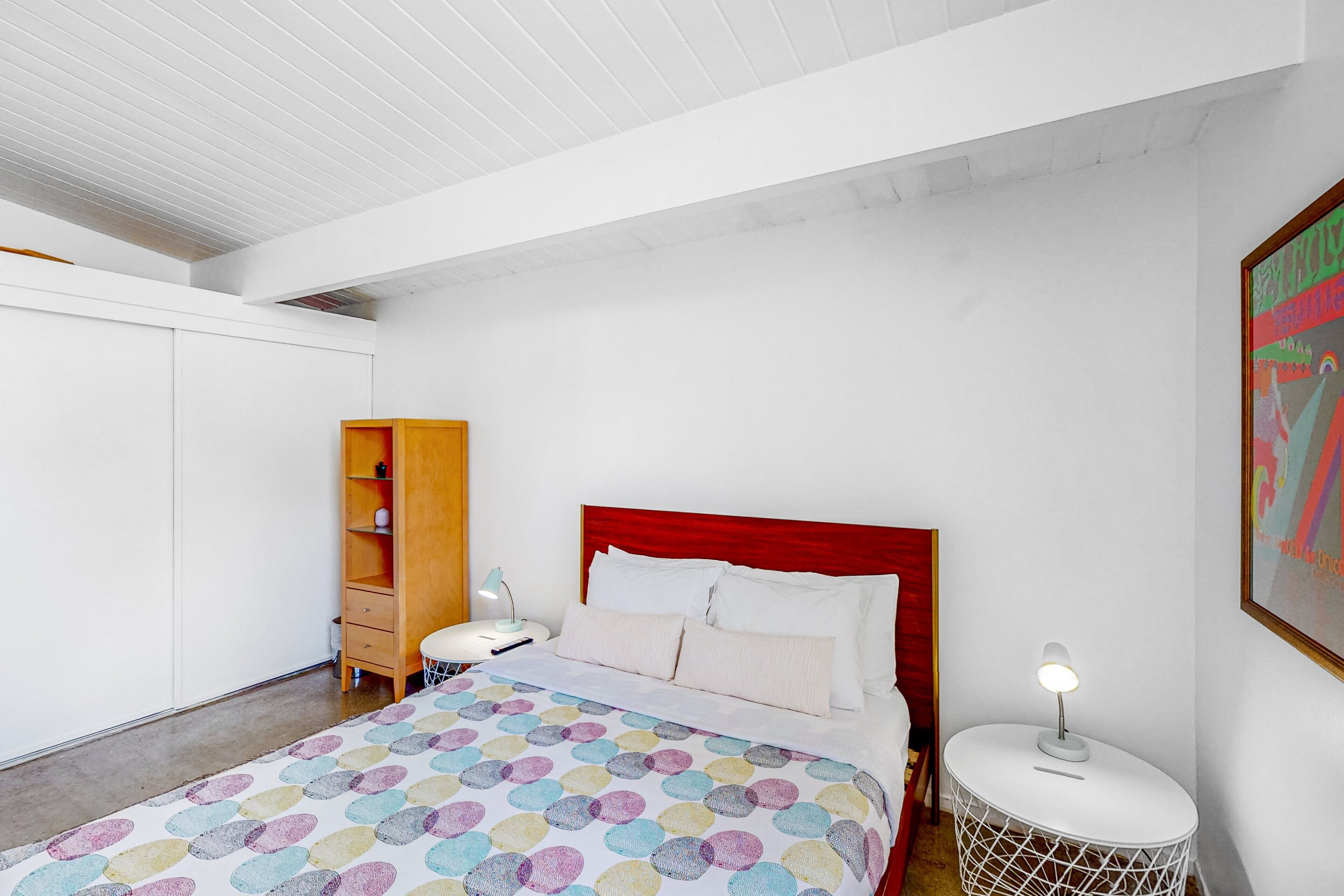 Ein Schlafzimmer mit einem Bett, einem hölzernen Nachttisch, einem weißen runden Beistelltisch, einem weißen Kleiderschrank und einem farbenfrohen, abstrakten Gemälde an der Wand.