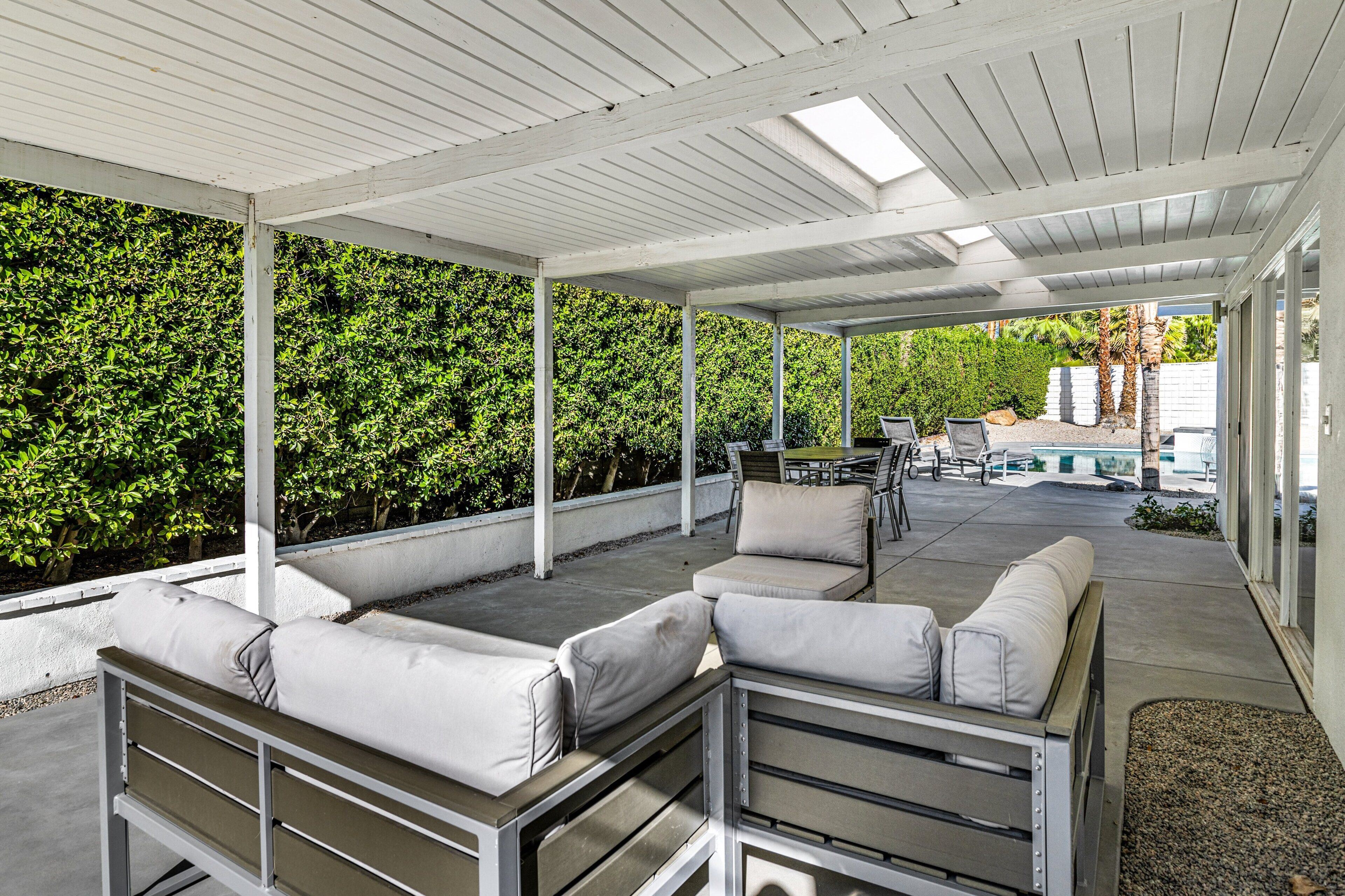 An outdoor patio with a covered seating area, a pool, and a lush green hedge.