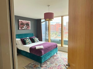 Ein modernes Schlafzimmer mit einem türkisblauen Bett, violetter Bettwäsche und Blick auf die Stadt durch große Fenster.