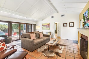 A spacious living area with a sofa, a coffee table, and a fireplace. There is a sliding glass door leading to an outdoor patio. - Charming desert getaway with lush garden, private pool & spa - close to downtown (Palm Springs)