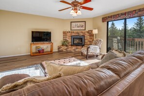 A living room with a leather couch, a fireplace, a television, and a balcony with a view of trees. - Placid home with new fireplace, full kitchen, gas grill, patio, & washer/dryer (Flagstaff)