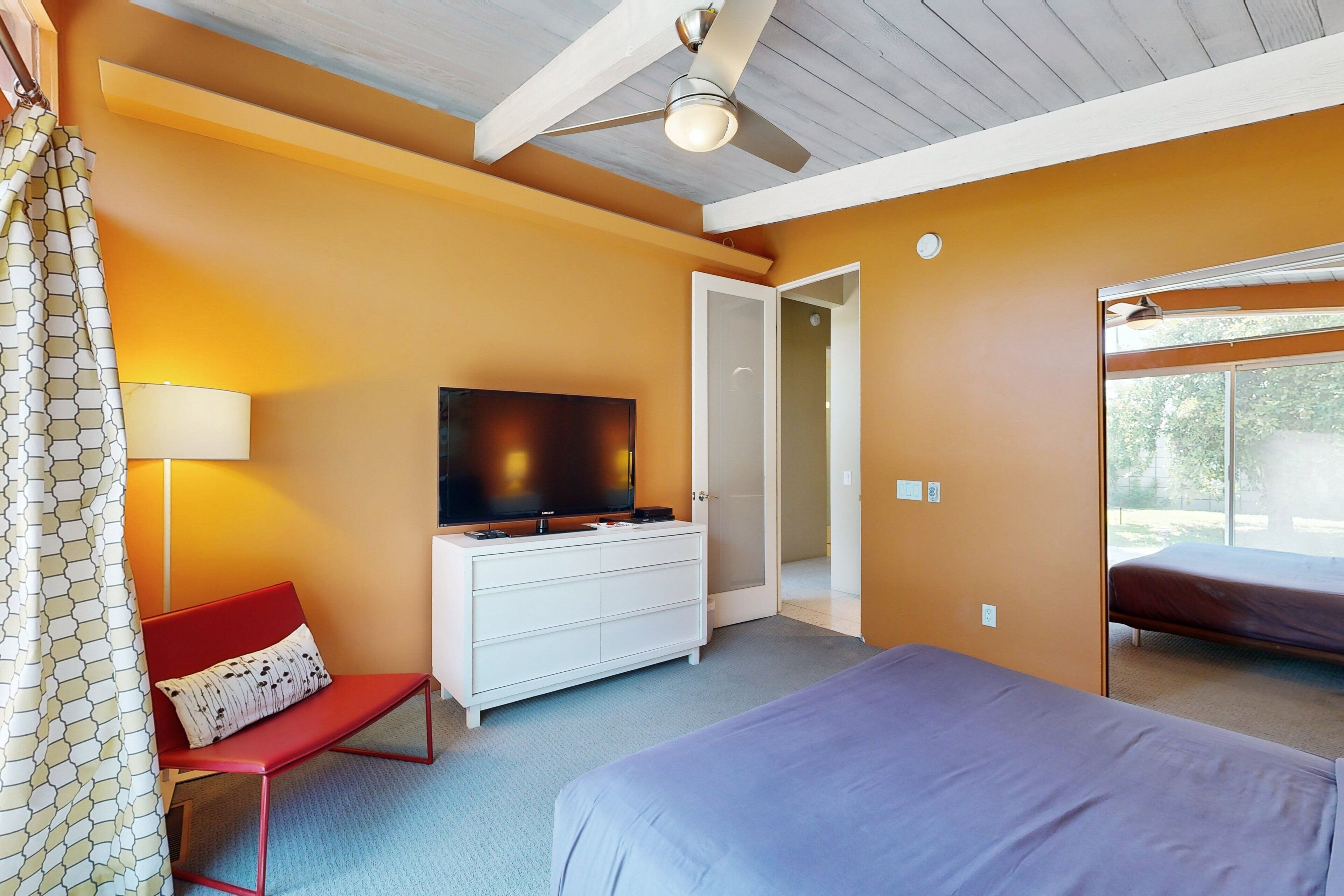 A hotel room with a bed, a TV on a white dresser, a red chair, and a ceiling fan.