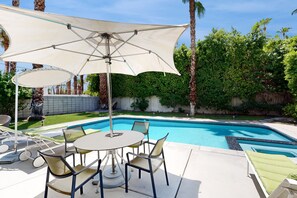 Un espace aménagé au bord de la piscine, avec une table et des chaises, un grand parasol et une piscine.