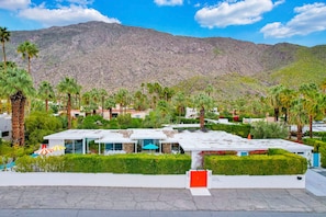 A modern hotel with a flat roof, surrounded by palm trees and a mountainous backdrop. - Mid-century modern estate with a pool, hot tub, gourmet kitchen, and fireplace (Palm Springs)