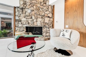 A modern living room with a stone fireplace, a glass coffee table, a red ottoman, and a patterned armchair. - Mid-century modern estate with a pool, hot tub, gourmet kitchen, and fireplace (Palm Springs)