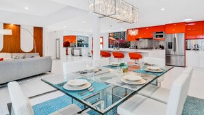 A modern kitchen with stainless steel appliances, a dining area with a glass table and white chairs, and a living area with a grey sofa and a wooden accent wall. - Mid-century modern estate with a pool, hot tub, gourmet kitchen, and fireplace (Palm Springs)