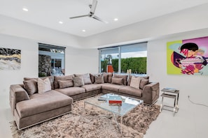 A modern living room with a sectional sofa, a glass coffee table, and a large window offering a view of greenery. - Mid-century modern estate with a pool, hot tub, gourmet kitchen, and fireplace (Palm Springs)