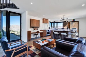 A modern kitchen with wooden cabinets, a dining area with bar stools, and a large window offering a view of the desert. - Desert stunner in Joshua Tree with mountain view - near town & park entrance (Joshua Tree)