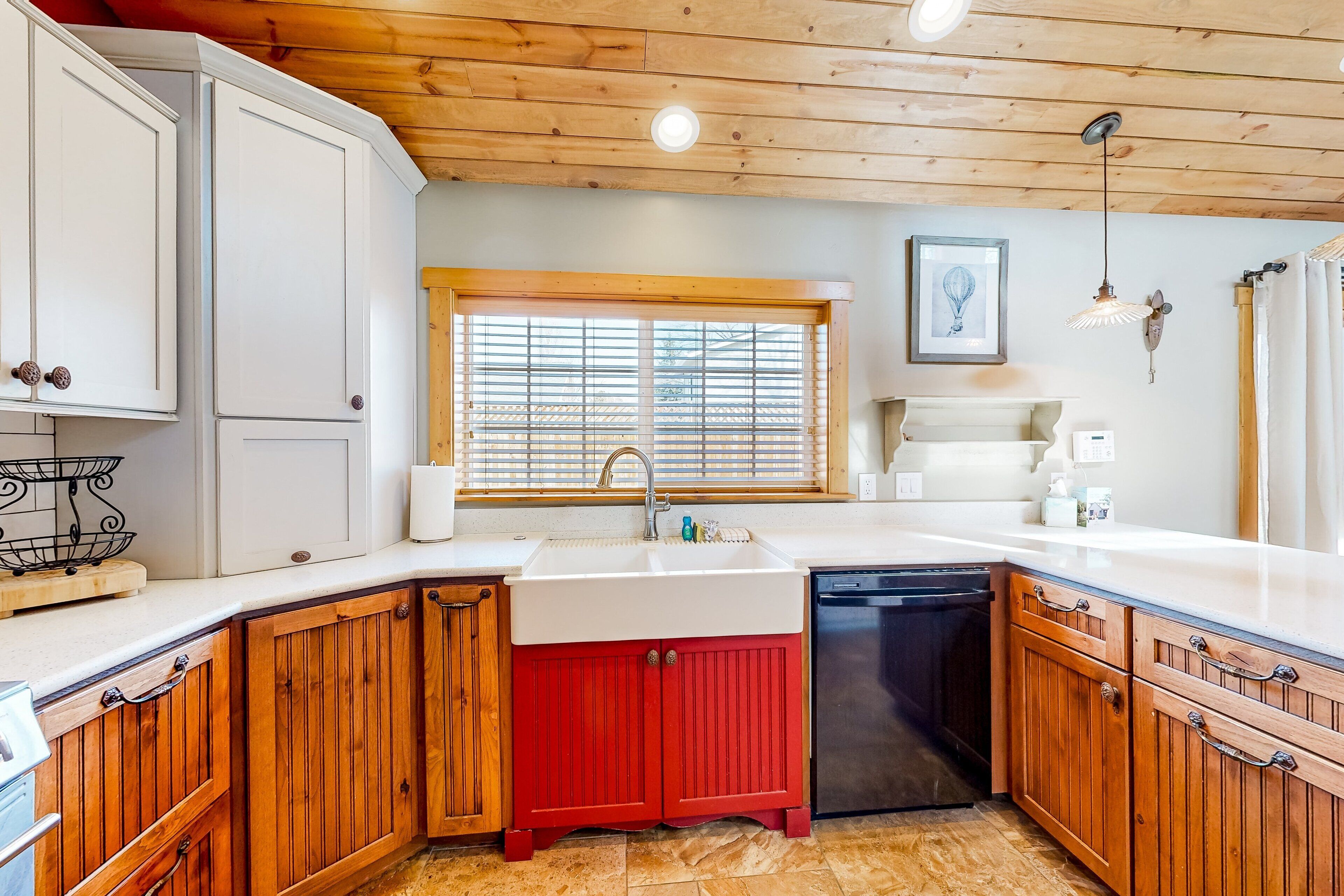 A kitchen with wooden cabinets, a red lower cabinet, a white countertop, a sink, and a window with blinds.