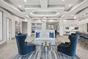 Eine moderne Hotelhalle mit einer Couch, zwei Sesseln, einem Couchtisch, einer Bar und einer Küche im Hintergrund.