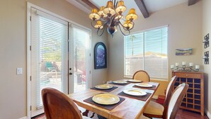 A dining area with a wooden table set for six, flanked by chairs with cushioned seats. A chandelier hangs above, and there are two windows with blinds. A small cabinet with a decorative top is to the right. - Elegant house with great outdoor space, private pool & spa - 3BR - #066636 (La Quinta)