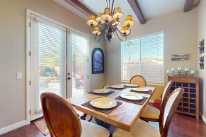 A dining area with a wooden table set for six, flanked by chairs with cushioned seats. A chandelier hangs above, and there are two windows with blinds. A small cabinet with a decorative top is to the right. - Elegant house with great outdoor space, private pool & spa - 3BR - #066636 (La Quinta)