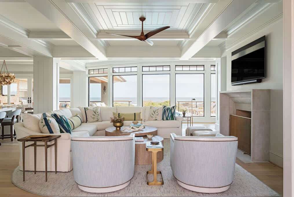 A spacious living room with a large sofa, two round side tables, a ceiling fan, and a TV mounted on the wall.