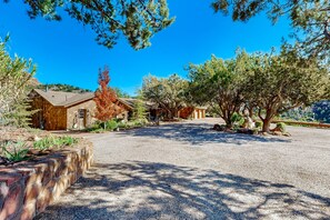 A spacious driveway leading to a stone building with a sloped roof, surrounded by mature trees and a clear blue sky. - Beautiful Sedona home with large country kitchen & amazing, 280-degree views (Sedona)