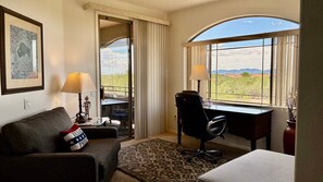 A hotel room with a desk, a chair, a sofa, a window with a view of the desert, and a framed picture on the wall. - Open & bright condo with WiFi, W/D, pool, hot tub & panoramic views (Oro Valley)
