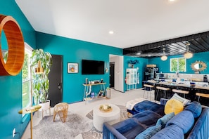 A modern living room with teal walls, a large TV, and a kitchen area with bar stools. - Dog-friendly home near Joshua Tree NP with Cowboy pool, mountain view, firepit (Joshua Tree)