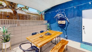 Eine Terrasse im Freien mit einem Holztisch und -stühlen, einer blauen Wand mit Wandgemälde und einem Holzzaun.