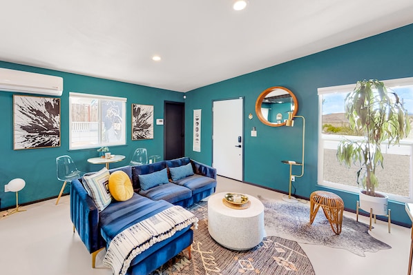 A modern living room with teal walls, a blue sectional sofa, a round coffee table, and a large window with a view. - Dog-friendly home near Joshua Tree NP with Cowboy pool, mountain view, firepit (Joshua Tree)