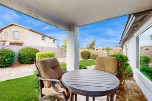A covered patio with a round table and two chairs, surrounded by greenery and a view of neighboring houses.