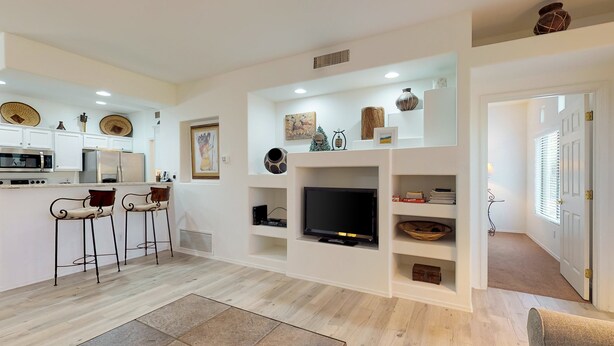 Een moderne keuken met een ingebouwde magnetron, koelkast en barstoelen. Een flatscreen televisie is aan de muur bevestigd boven een plank met decoratieve objecten.