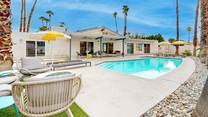 Un área de piscina con sillones de descanso y sombrillas, rodeada de palmeras y un edificio con varias habitaciones.
