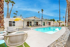 A pool area with lounge chairs and umbrellas, surrounded by palm trees and a building with multiple rooms. - Mid-century modern hacienda with pool, outdoor kitchen, & fireplace - dogs ok (Palm Springs)