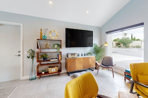 A modern living room with a flat-screen TV, a wooden cabinet, and a large window offering a view of the outdoors. - Mid-century modern hacienda with pool, outdoor kitchen, & fireplace - dogs ok (Palm Springs)