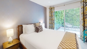 Habitación de hotel con cama, mesita de noche con lámpara, ventilador de techo y vistas a un jardín a través de una puerta de vidrio.